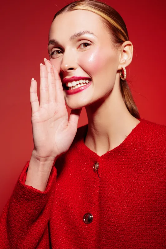 Smiling woman in red sweater whispers with hand near mouth on red background. Concept of communication, secret, happiness, and positive emotion in studio portrait.