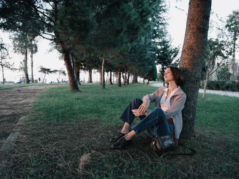 A calm woman sits beneath a tall tree in a tranquil park, taking a moment to reflect. She wears casual clothes and emits thoughtful serenity, creating a peaceful outdoor mood.
