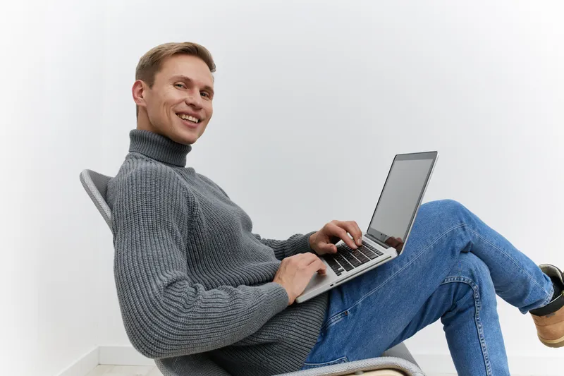 Smiling man working on a silver laptop while sitting on a chair isolated on plain white background. Young casual male holding computer device, happy expression, technology, indoors, lifestyle concept,