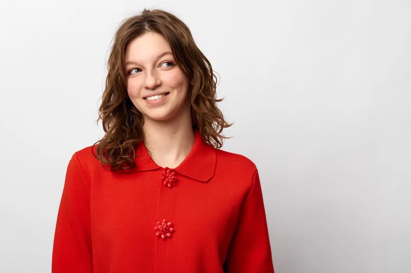 Young woman with curly brown hair wearing a red sweater smiles softly looking to the side on a white background. Concept of casual style, happiness, and natural beauty.