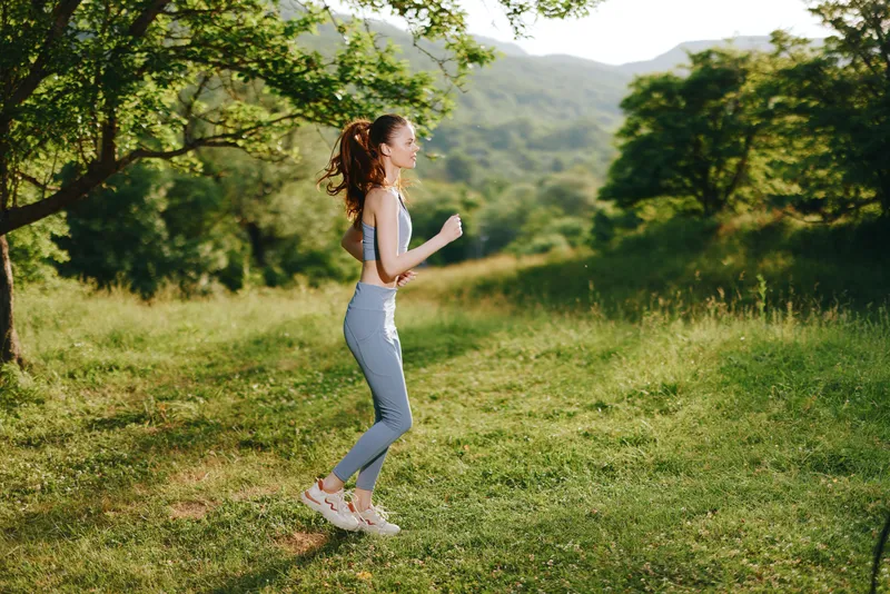 Fit young woman jogging in a green landscape during summer, wearing activewear, exuding happiness and vitality, illustrating a healthy lifestyle