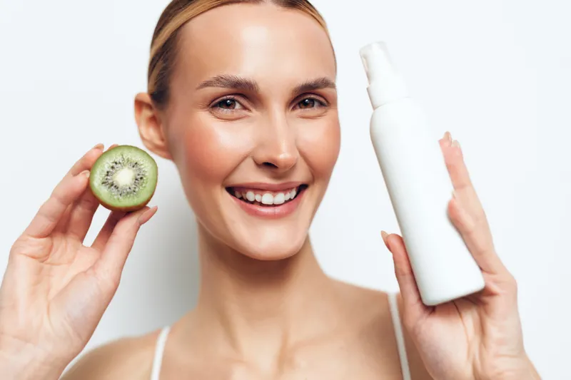 Smiling woman in beauty skincare, holding kiwi slice and cream bottle, showing serum concept, healthy glow on face, clean white studio, soft light