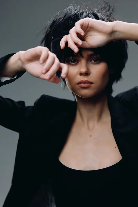 Portrait of a serious woman with dark hair wearing a black outfit, posing with hands near face against a grey background.