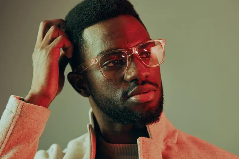 Stylish young African man in glasses wearing modern casual attire, looking thoughtfully with a bold red and green color palette in a minimalist background