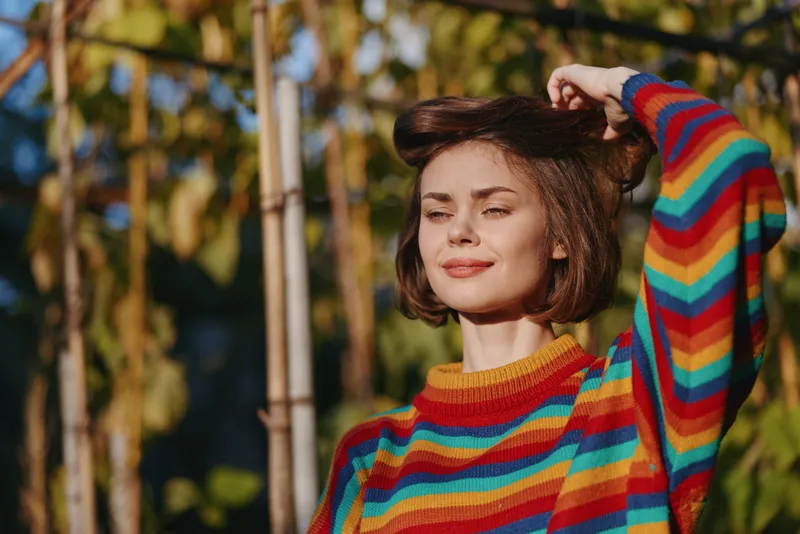 Woman portrait in colorful striped sweater smiling outdoors in autumn sunlight, short hair and relaxed expression. Casual fashion, warm tones and natural background for lifestyle mood.
