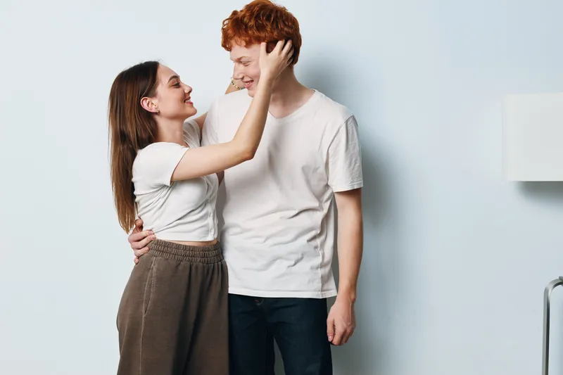 Couple, young woman and man embracing with smile, standing close and looking at each other in love and happiness at home with white wall background.