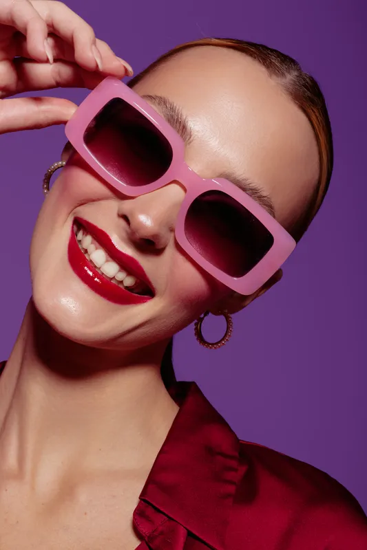 Smiling woman with pink sunglasses and red lipstick poses against purple background. Fashion portrait with bright makeup and stylish accessories for beauty and glamour concept.