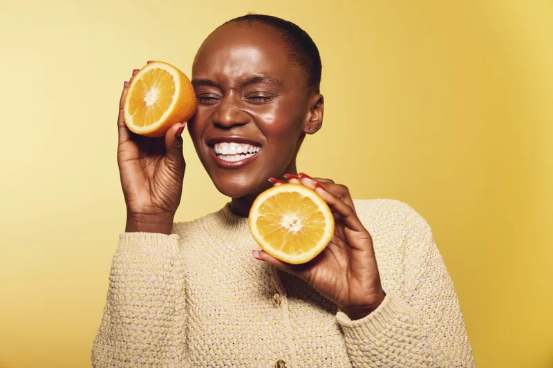 Smiling dark skinned African woman holds two orange halves near face against yellow background. Concept of fresh fruit, healthy lifestyle, vitamin, and natural beauty with joyful expression.