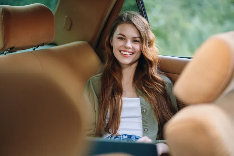 Young woman smiling in a car, wearing a casual outfit with natural beauty, enjoying a summer day, surrounded by soft colors and a serene green background.