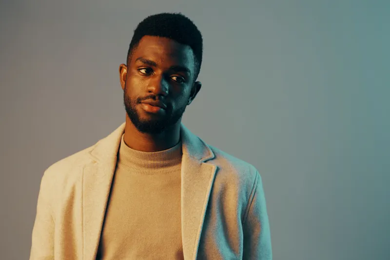 Elegant young Black man in a stylish gray blazer and cream sweater against a soft blue background, expressing a reflective mood