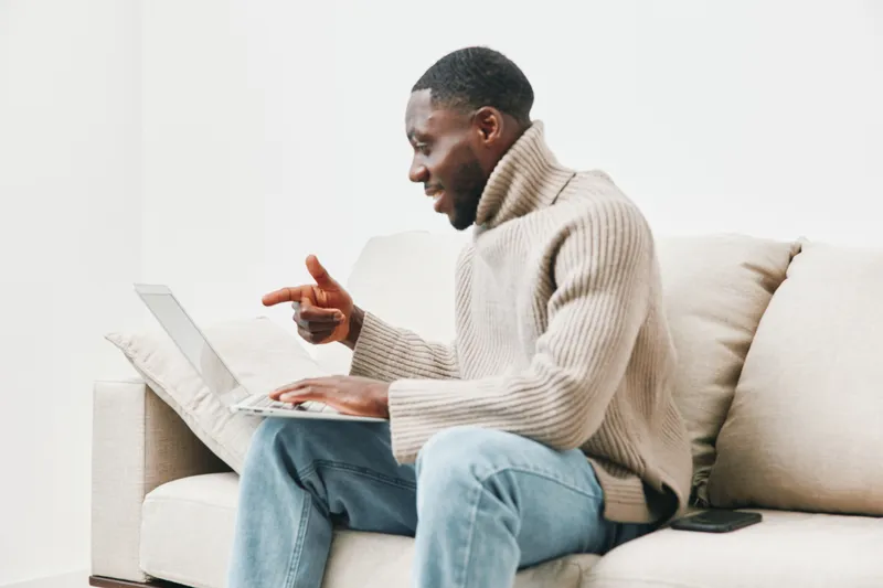 Young Black man in a cozy sweater joyfully working on a laptop at home, showcasing a relaxed atmosphere and modern interior design