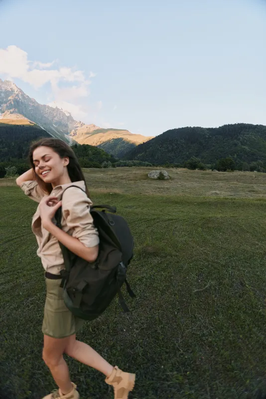 Smiling woman with a backpack walking through a meadow near mountains, showcasing outdoor lifestyle, exploration, wellness, and carefree travel moments.