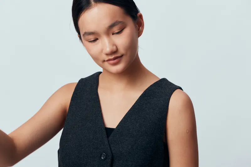 Young woman with closed eyes wearing dark sleeveless vest standing against light gray background in studio. Asian female model with natural makeup and calm expression portrait with minimal style
