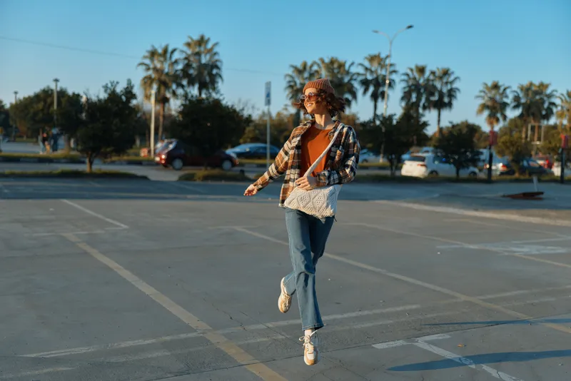 woman jumping in parking lot by palm trees with casual bag and smile, sunlight golden hour glow creates authenticity and candid lifestyle mood, mindful living and emotional storytelling