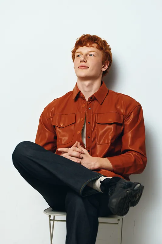 Young man with red hair and casual orange shirt sitting crosslegged on chair against white background looking to side thoughtful confident relaxed posture.