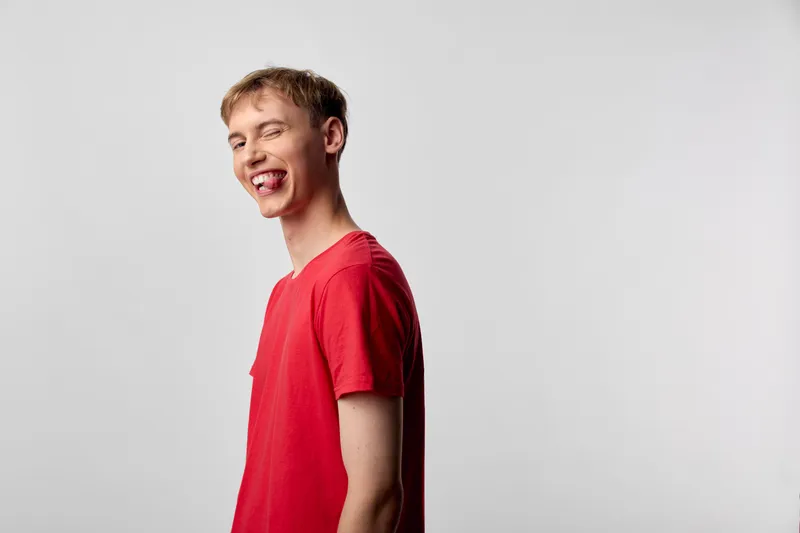Playful young man in a red shirt sticks out his tongue against a plain backdrop, casual candid mood, energetic expression, humor and lighthearted vibe for lifestyle campaigns and youth messaging