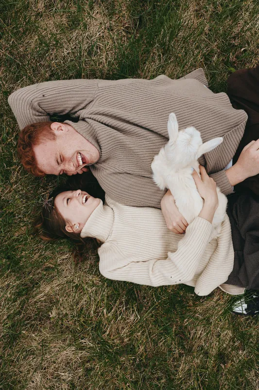 Young couple lies on grass holding a white baby goat while smiling and enjoying a relaxed lifestyle moment outdoors in cozy sweaters during autumn.