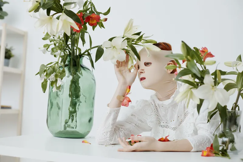 Ethereal portrait of a model in a white dress surrounded by lilies, showcasing emotions of contemplation and tranquility in a light, minimalist setting