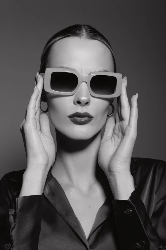 Fashionable white woman with pale skin wears large sunglasses and holds them with both hands against plain dark background. Concept of style and elegance.