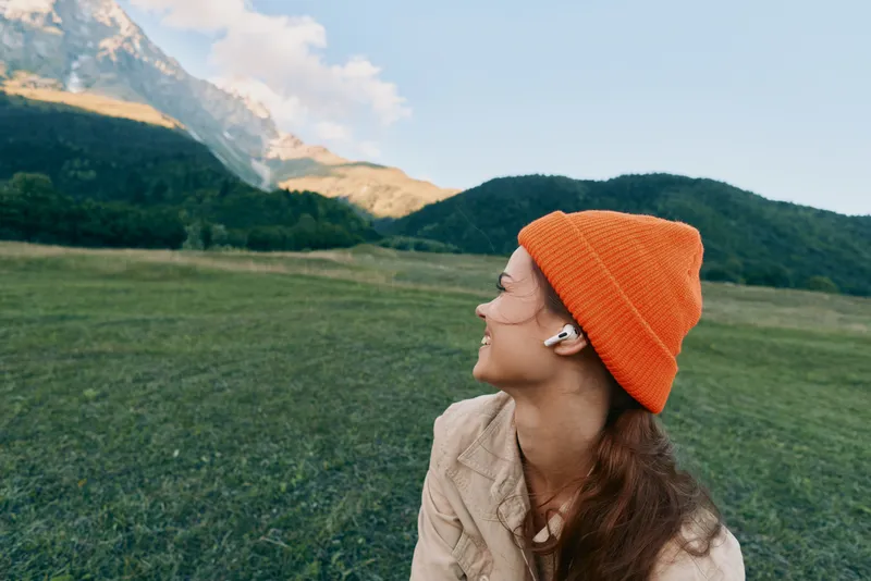 Woman outdoors, side profile, orange beanie, headphones on, gazing at distant mountains over a green field, serene lifestyle moment in natural light and fresh air.