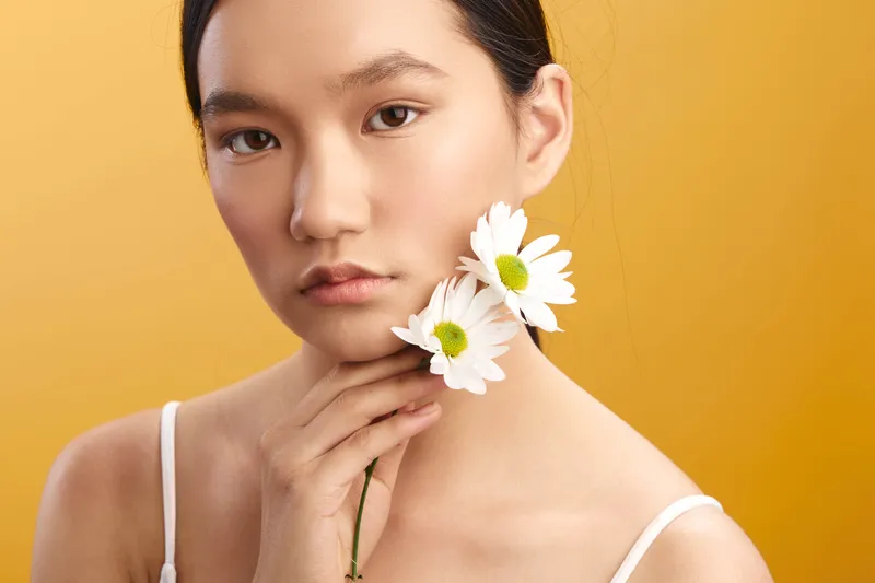 asian woman gazes softly at the camera, holding daisies near her chin, a serene portrait with gentle lighting, calm expression, and a minimalist warm background.