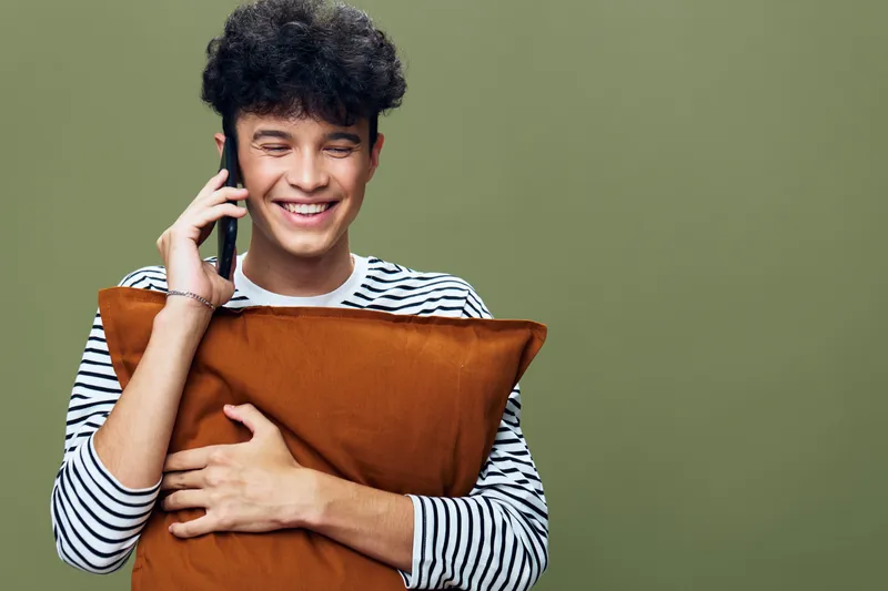 Smiling man wearing striped shirt hugging orange pillow while talking on phone against green background, showing joy and casual style in indoor setting.