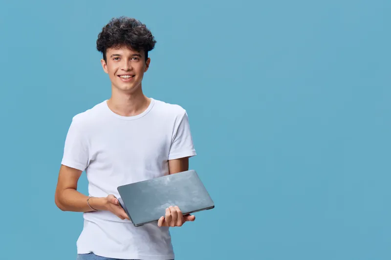 smiling young man holding laptop with casual white t-shirt, cheerful expression, standing on blue background in studio lighting