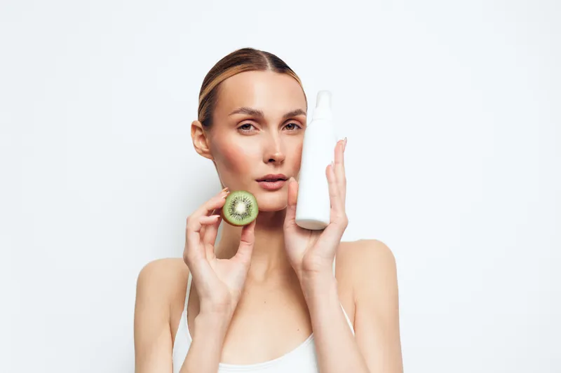 Young woman holds fresh kiwi and white cosmetic bottle near face shows natural skincare concept with clean fresh healthy skin isolated on white background.