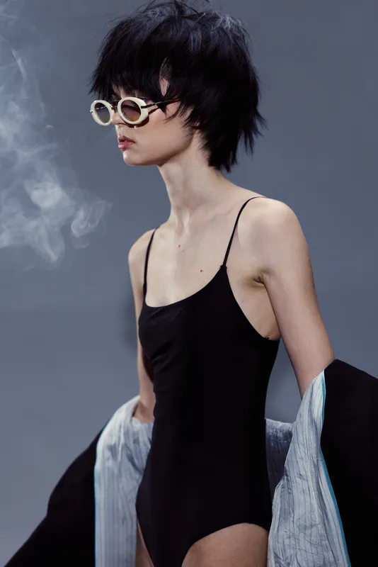 Slim woman wearing black swimsuit and sunglasses posing with jacket on gray background with smoke