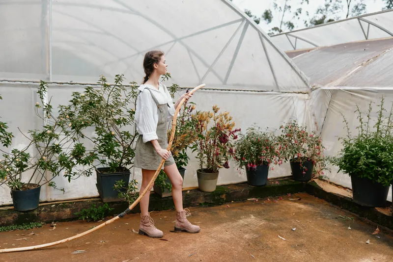Young woman gardening in a greenhouse, wearing a casual outfit and boots, surrounded by blooming plants and flowers, embodying a nurturing and vibrant atmosphere