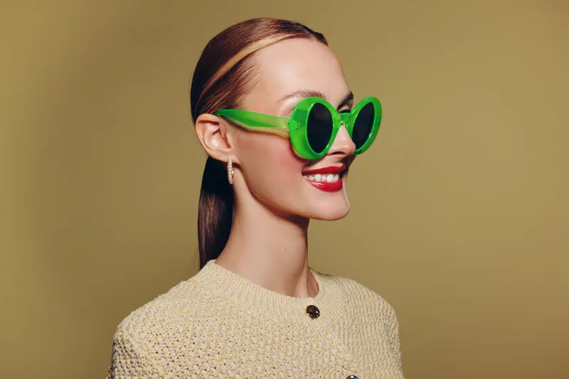 Smiling woman with slicked hair wears large green sunglasses and beige knitted sweater. Studio portrait on yellow background. Concept of fashion, style, and confident female model.