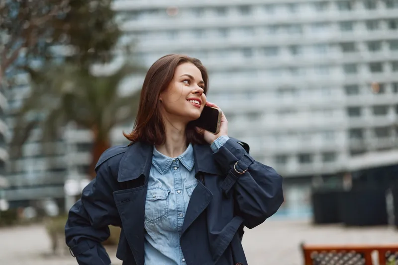 A smiling woman in a denim shirt and dark jacket chats on her phone outdoors, with a modern building backdrop and blue skies, conveying confident urban style and everyday professionalism.