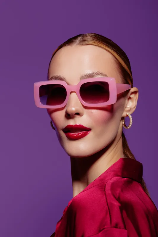 Stylish woman with pink sunglasses and red lipstick poses on purple background. Fashion portrait with trendy accessories and vibrant colors. Concept of glamour, beauty, and modern style.