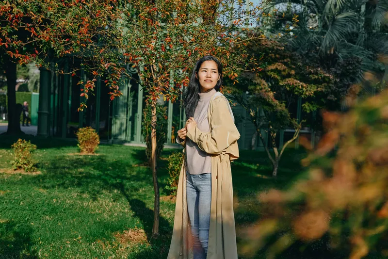 Woman wearing a beige trench coat stands in a sunny garden among trees and blooming plants, showcasing outdoor fashion, casual lifestyle, and a relaxed pose.