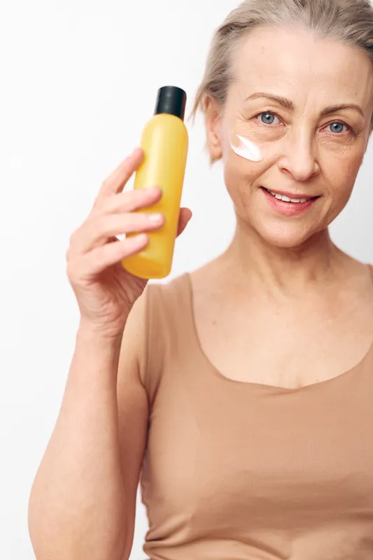 Mature woman with light skin applying cream on cheek holding yellow bottle. Bright studio background. Concept of skincare, aging care, beauty routine, and healthy lifestyle.