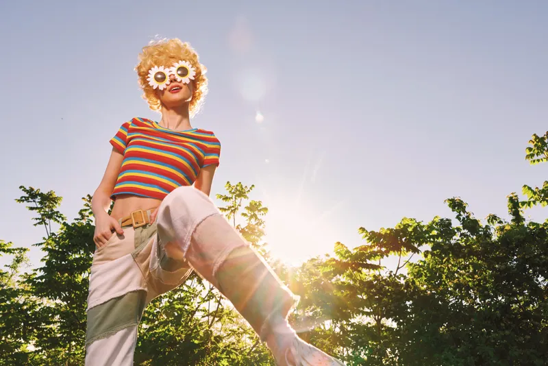 Woman outdoors in bright daylight wears a striped shirt and retro sunglasses, posing playfully with a flower crown, capturing a carefree summer mood and vibrant color aesthetic.