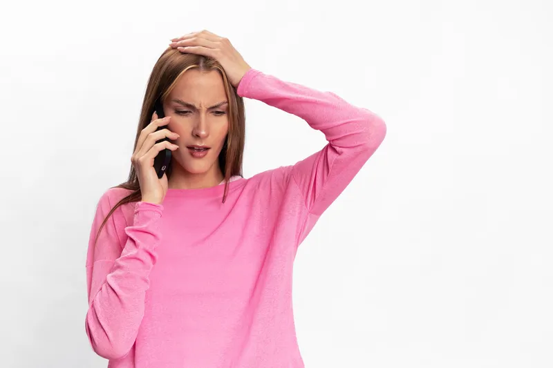 Young woman in pink sweater talks on phone with worried expression holding head. Isolated on white background. Concept of stress, confusion, and difficult phone conversation.