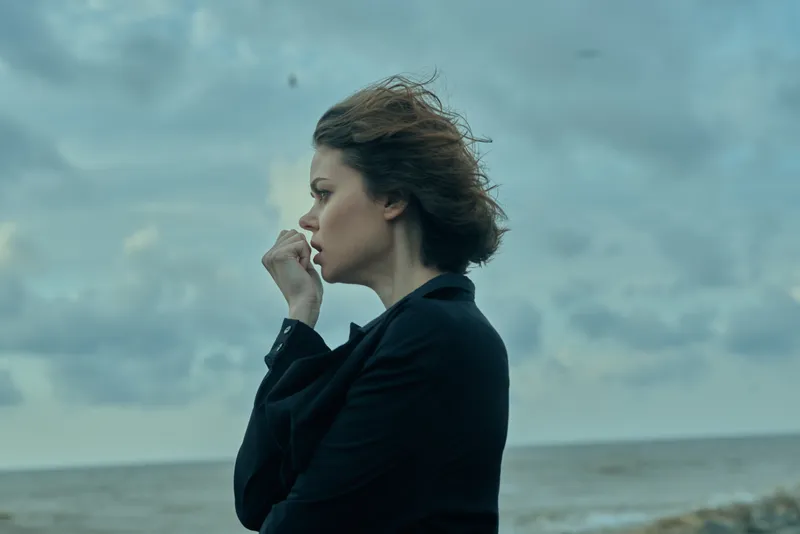 Thoughtful woman in a dark blazer by the sea, wind in hair, standing on a windy coastline, conveying introspection, resilience, and calm amid ocean exposure.