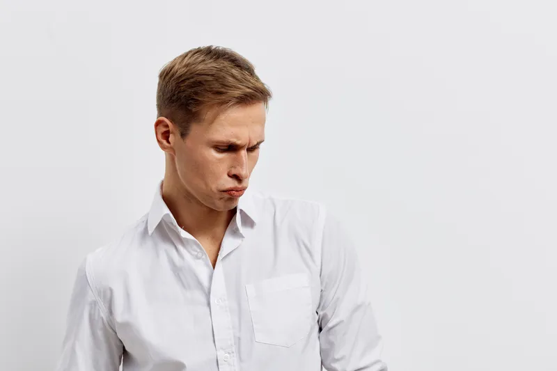 Young man with short brown hair wearing a white shirt looks down with a serious expression, against a plain white background in studio portrait, minimalistic, professional concept.