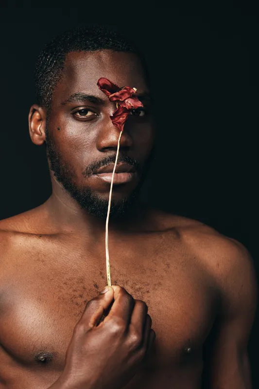 African man with dark skin holding dried flower in front of face, creative conceptual portrait with visual style and artistic expression on black background.