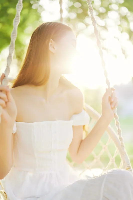 Young woman enjoying a sunny day on a swing, wearing a white dress, with soft sunlight illuminating her face, capturing a feeling of serenity and joy