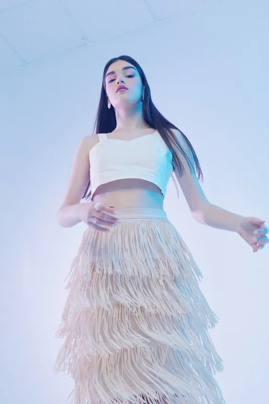 Young woman in a white crop top and fringed skirt poses confidently in a studio with soft lighting. Concept of woman beauty fashion and modern style expression.