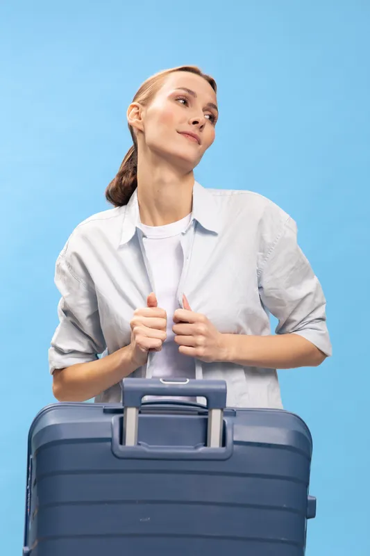 Young woman in casual clothes holds a blue suitcase and looks away on light blue background. Concept of travel, vacation, journey, and summer holidays preparation.