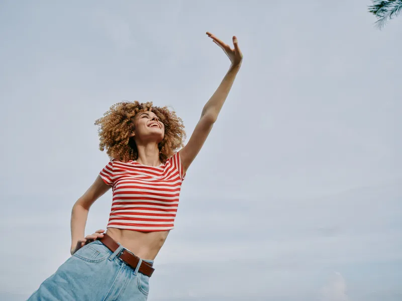 Young woman with curly hair wearing a red striped crop top and denim jeans raises an arm in a joyful pose against a clear blue sky.