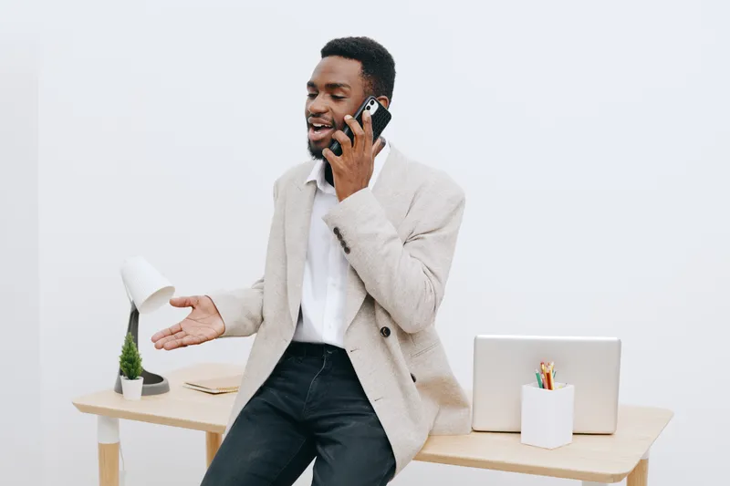 Smiling man talking on phone in office setting with desk, laptop, and decor, casual professional attire, cheerful mood, modern workspace, neutral background, indoor business communication concept, lif