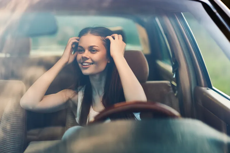 Young woman with long hair smiling inside a car, enjoying a sunny day, reflecting joy and freedom in a natural setting.