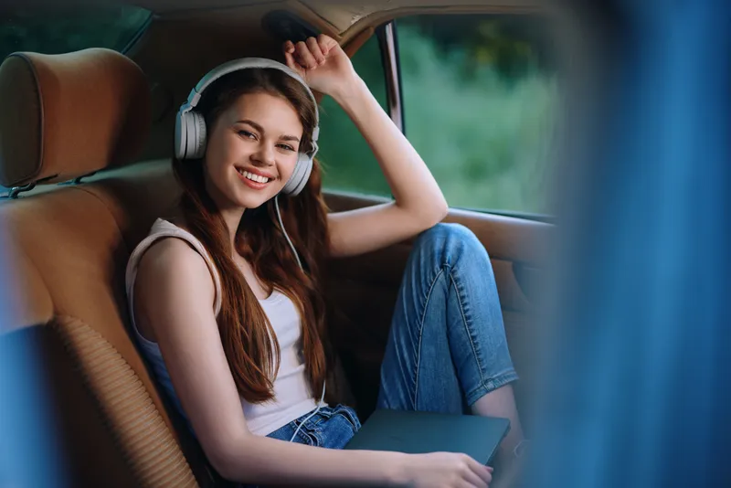 Happy young woman wearing headphones in a car, enjoying music and relaxation, with a green outdoor background. Ideal for travel and lifestyle themes.