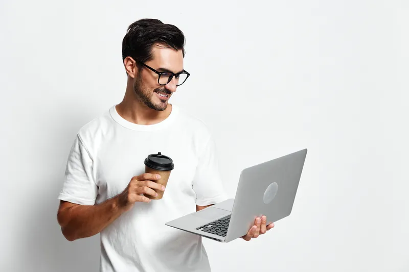 Smiling man holding coffee cup while using laptop, wearing casual white t-shirt with glasses, standing against plain light background. Work lifestyle concept.