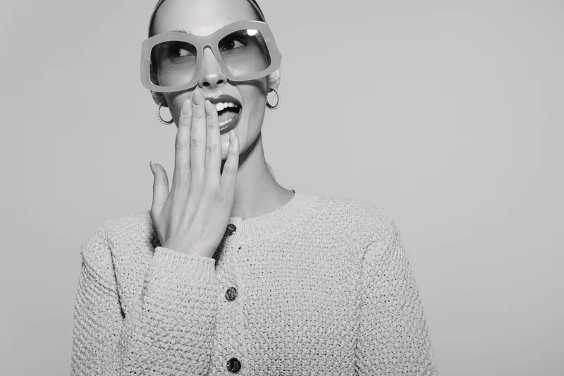 Happy Caucasian woman with fair skin and oversized sunglasses laughs, hand near mouth. Bright studio portrait in monochrome style.