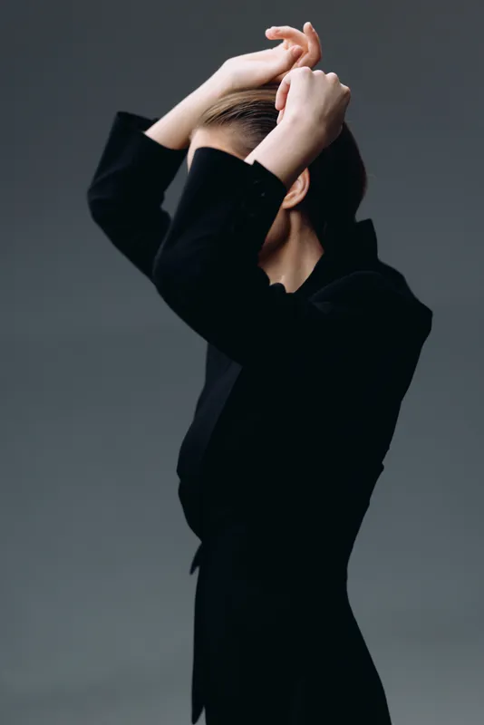 Elegant woman in black outfit posing with hands on head against gray background.
